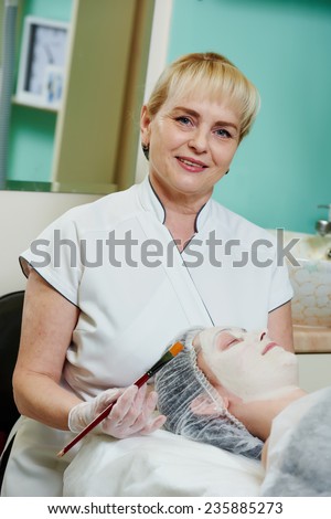 beautician worker with paintbrush applying facial cosmetic mask to female client in beauty salon