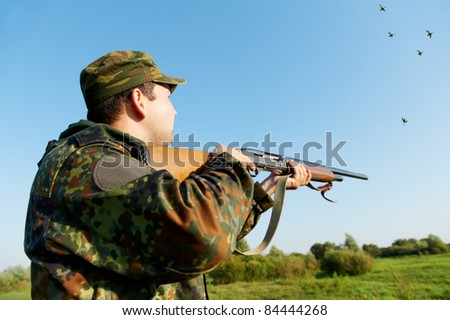 Male hunter in camouflage clothes on the field aiming and shooting with hunting rifle to gamebird during a hunt