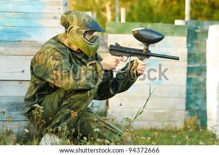 Happy paintball sport player man in protective camouflage uniform and mask with marker gun outdoors