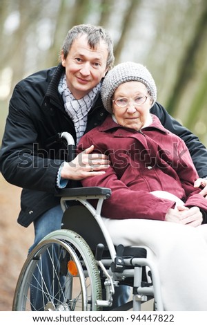 Caregiver careful man walking and embracing disabled senior woman grandmother at wheelchair in nature