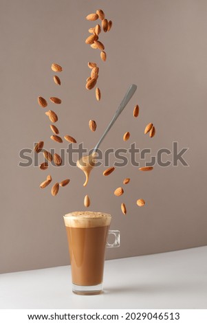 Glass cup with cappuccino and fragrant foam and a spoon and falling almonds. Cup with cappuccino, latte or coffee with milk. Creative design for cafes and restaurants with a cup of coffee. 
