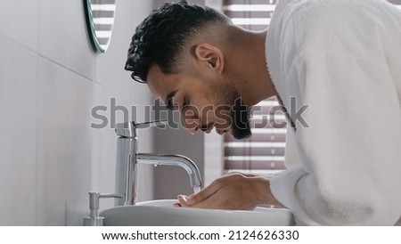 Side view arab spaniard indian arab man guy opens faucet near washbasin at home in hotel bathroom washes face with hot cold water morning beauty ritual male hygiene guy in bathrobe wet facial wash Stock fotó © 