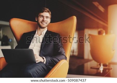 Young smiling successful man entrepreneur in formal business suite with a beard sitting on orange armchair with laptop in luxury office interior ストックフォト © 