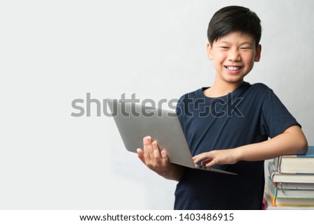 Online learning concept. A smart looking Asian boy standing with a computer laptop and a tall stack of colorful and various types of text books beside him. E-learning, Gamification, Self- Study. 