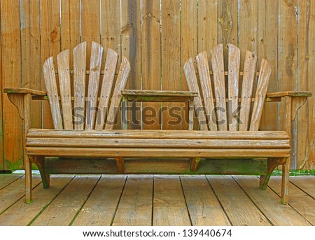 A close up of a dual wooden seat with a center table on a wooden deck/patio with a fence behind it for privacy