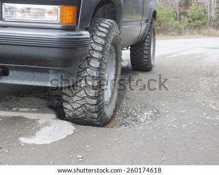 A black SUV with a front wheel sunk in a water filled pothole on an old rural asphalt/dirt road