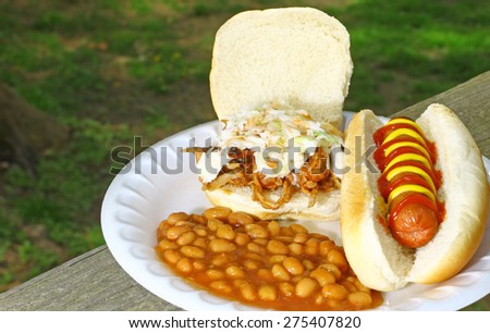 A fresh made BBQ sandwich with coleslaw and hot sauce along with a grilled hotdog in a bun with catchup & mustard and a side of home made baked beans served on a Styrofoam plate outside on a table