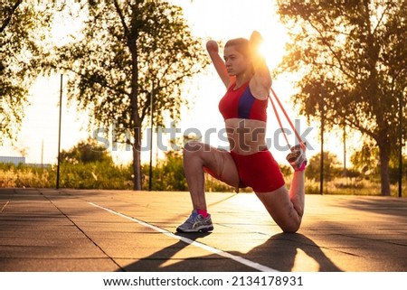 Young woman athlete with an amputated arm and burns on her body limber up on the sports field. Resistance band exercise outdoor at sunset Imagine de stoc © 