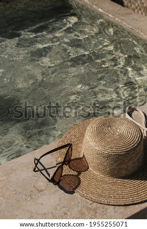 Sunglasses and straw hat on marble swimming pool side with clear blue water with waves sunlight shadow reflections. Minimal aesthetic summer vacation concept background.