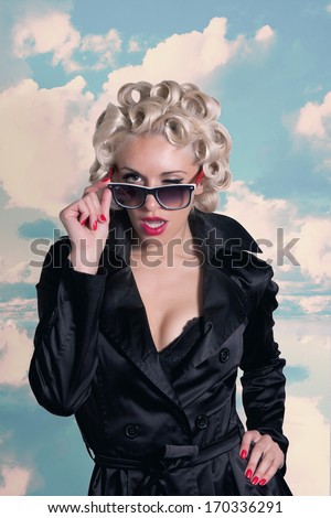 beautiful young woman  on sky background in old fashion clothes representing pinup and retro style winks. Girl dressed in a black cloak with red lipstick and sunglasses.