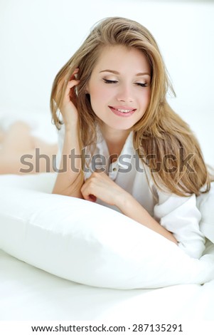 A woman lying at the end of the bed underneath the quilt and smiling, with her head resting upon her hand with the other in her hair.