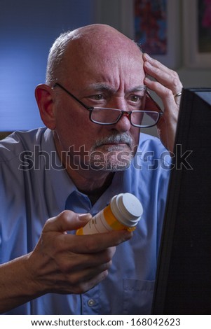 Confused older man at computer and holding prescription bottle, vertical