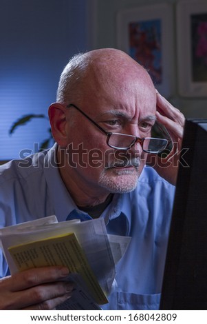 Distressed older man at computer and holding bills, vertical
