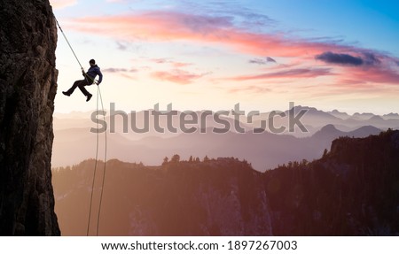 Adventurous Man Rappelling from Cliff. Aerial view of the mountains during a colorful and vibrant sunset or sunrise. Landscape taken in British Columbia, Canada. composite. Concept: Adventure Сток-фото © 