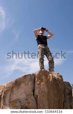 The man at top of a rock on background sky and looks into binoculars