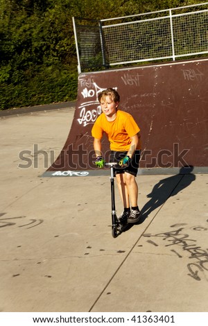child with scooter and orange T-shirt is doing tricks at the scateboard parc and enjoying it