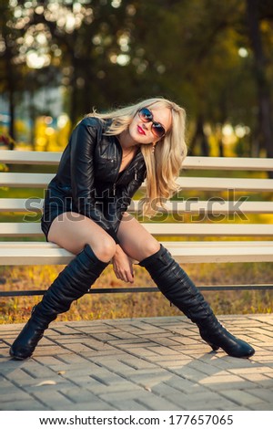 Sexy Girl in leather dress sitting on a bench in the park. Beautiful girl with lovely smile sitting on bench in park. Attractive sexy girl sitting on a park bench