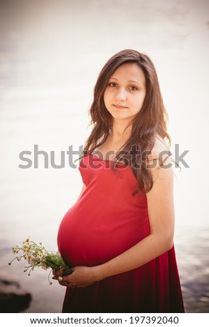 The young pregnant woman in red dress with a bouquet of wild daisies walks in the park by the lake. Riverside. Young pregnant woman walking on the beach