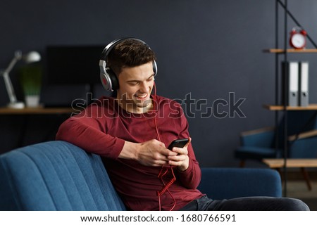 Enjoy listening to music.Young man in headphones listening music on smart phone using music app. Portrait of guy in earphones and mobile phone at home. Relaxation, leisure and stress management.