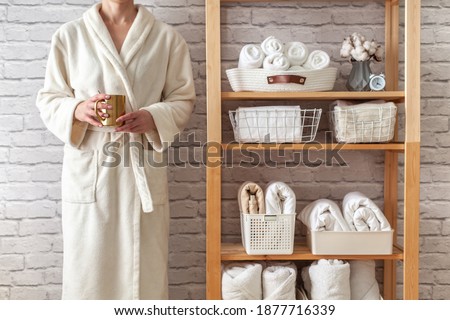 Woman in robe is standing and holding white wire basket of folded bed sheets near organized linen closet in bathroom. Sorted and folded towels in white wicker and wire baskets placed on wooden shelves