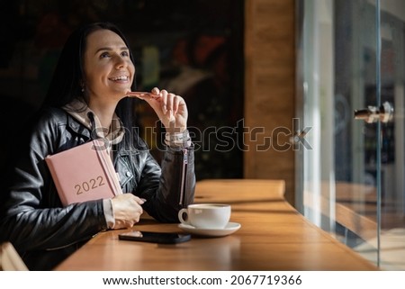 Inspired young woman with a smile looking through window and holding coral colored diary 2022. Hope and inspiration concept. Lady is smiling and dreaming about future new year. Happiness and success.