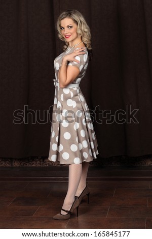 Cheerful girl in a brown retro-dress with polka dots on the background of dark-brown curtain