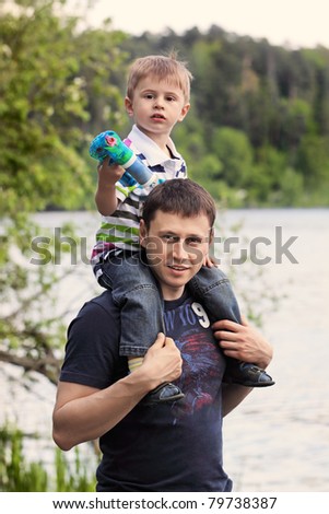 The little boy sits on the shoulders of the father near the lake in the park