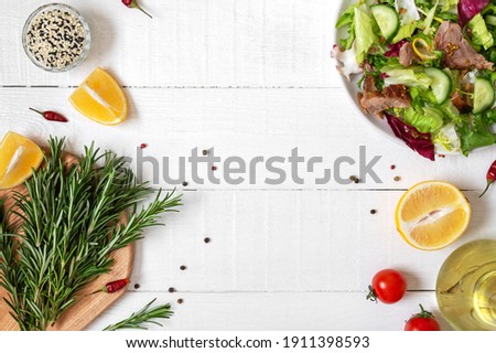Bowl of fresh salad, rosemary, lemon, spices and oil on white wooden table. Food background. Ingredients for cooking with copy space. Healthy eating concept.
