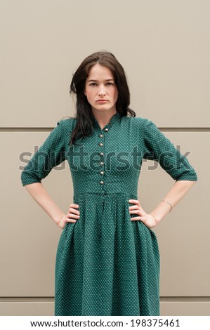 Young female model with natural look and hands on her hips, looking at the camera in front of a metal wall. She wears a green summer dress with white dots