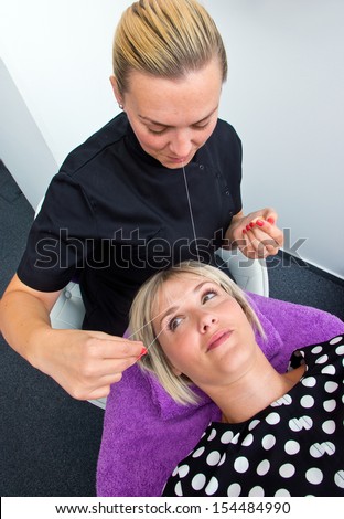 beautician makes threading hair removal procedure to blond woman in salon