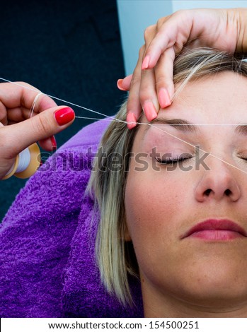 beautician makes threading hair removal procedure to blond woman in salon