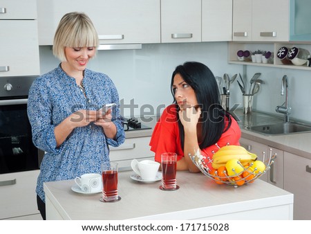 two attractive woman friends in kitchen , one bored other using mobile phone