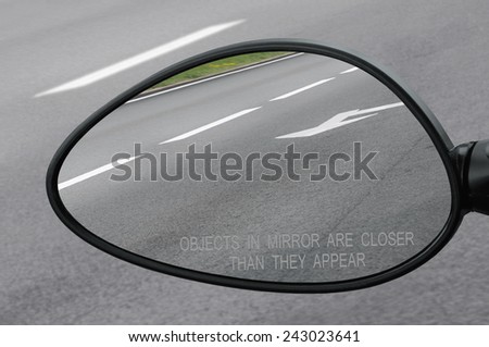 Rear view mirror with warning text objects in mirror are closer than they appear, reflecting road, left side lateral, macro closeup, tarmac asphalt background reflection, white lines, arrows marking