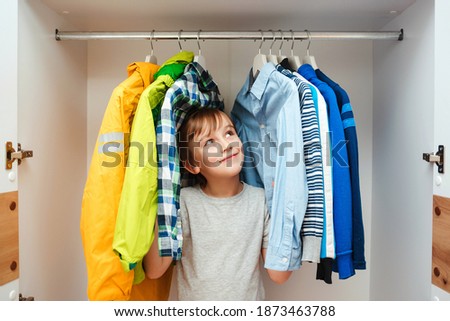 Happy smiling boy searching for clothing in a closet. Preteen boy chooses clothes in the wardrobe closet at home. Kid hiding among clothes in wardrobe.