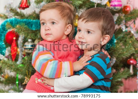 A cheerful toddler is hugging a little girl at the decorated New Year tree