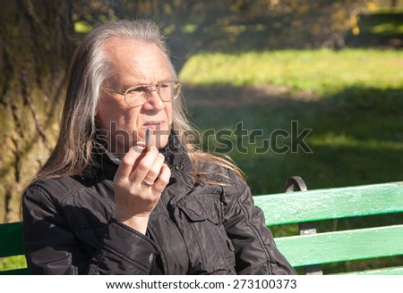 portrait of elderly gray-haired man in glasses, black jacket smoking a cigarette sitting on a bench in city park on sunny spring day closeup