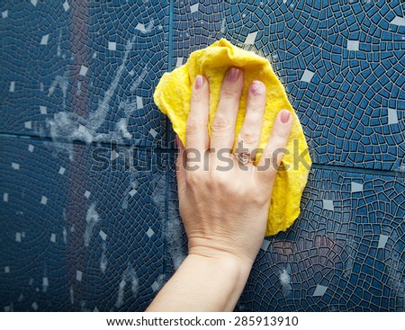 female hand washes the blue tile on the wall with a yellow cloth lather in the bathroom closeup