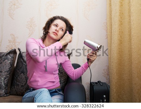 young brunette woman in pink blouse and blue jeans dries the hair dryer sitting on the couch