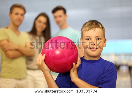 Smiling boy dressed in blue T-shirt holds pink ball in bowling club; two men and woman stand behind him; focus on boy; shallow depth of field