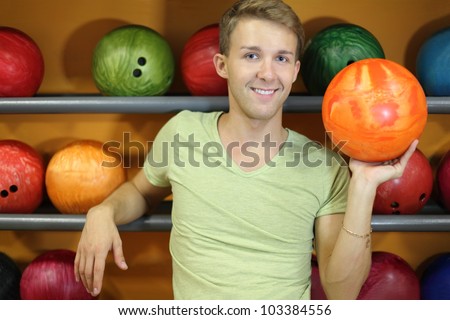 Happy man stands near shelves with balls and holds orange ball in bowling club; shallow depth of field