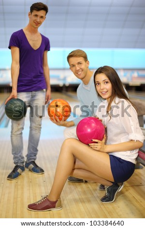 Two young men and girl with balls in bowling club; left man stands and others sit; shallow depth of field; focus on woman