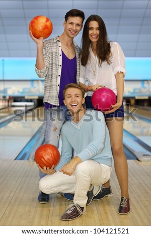 Two young men and beautiful girl hold balls in bowling club; focus on sitting man; shallow depth of field; full body