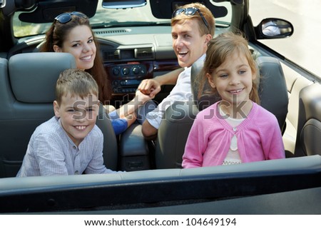 Young couple sits in cabriolet on front seats looking back, two children sit backwards on backseats, everybody laugh, back view