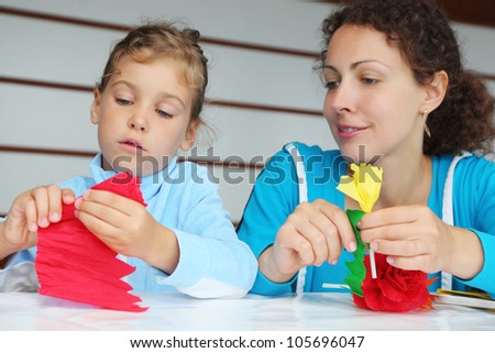 Mother and daughter sit at table and make artificial flower of tissue paper; focus on girl