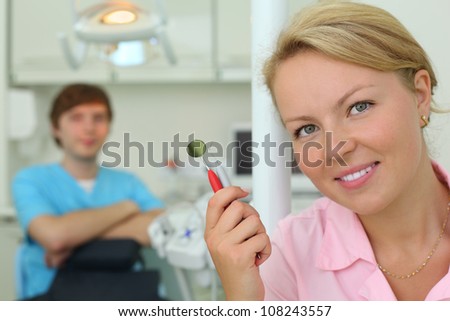 Smiling dentist with tools in cabinet of dental clinic; other doctor sits behind her. Focus on woman.