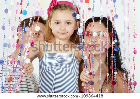 Little girl in striped sundress looks out from behind curtain of plastic beads, her parents stand behind curtain