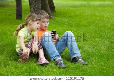 Boy and his younger sister sit on grass under tree in park and play electronic game