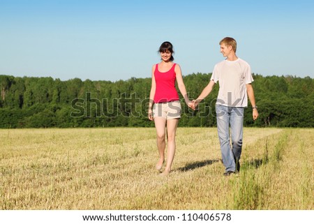Husband, wife holding hands walk in field near wood