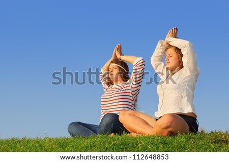 Two young girls meditate at green grass at background of blue sky.