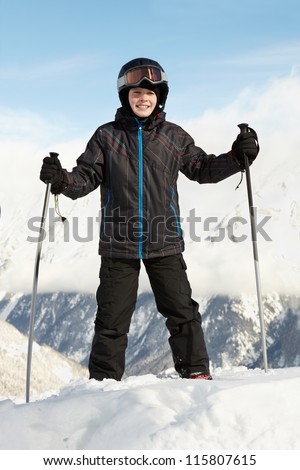 Boy in ski suit stands leaning on ski poles with blue sky and mountains on background